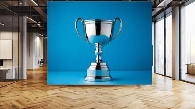 Shiny silver trophy with an elegant base, standing against a blue background. The cup is adorned with intricate designs and features two hands holding the handle at its top. It has been decorated. Wall mural