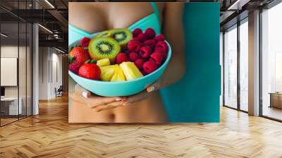 Closeup of a woman s hands holding a colorful bowl filled with fresh juicy fruits like kiwi strawberries raspberries banana and pineapple against a vibrant turquoise background  Healthy nutritious Wall mural