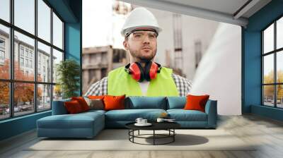 serious engineer in protective goggles and hardhat standing on construction site Wall mural