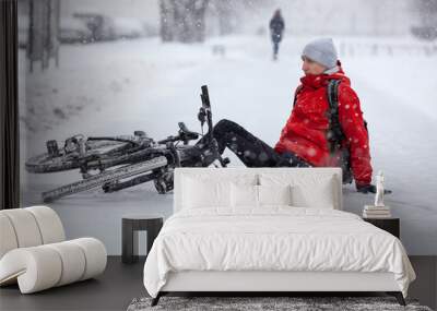 Fallen down Caucasian cyclist sitting on slippery roadside, riding bicycle in city at winter season Wall mural