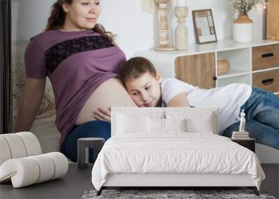 Happy little girl hugging pregnant mother belly while laying on the bed in bedroom Wall mural