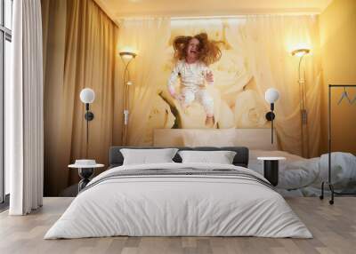 Cute little girl jumping on white bed in evening in sleeping room or in hotel Wall mural