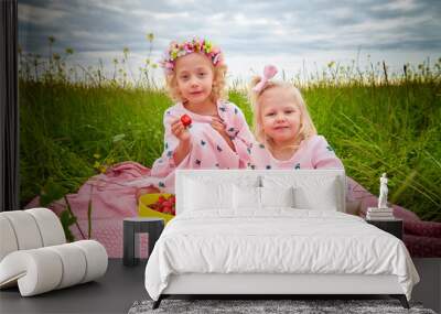 Two pretty young small blonde girls with curly hair in pink dress in field with grass and flowers in summer day. Sisters in photoshoot and cloudy sky background Wall mural