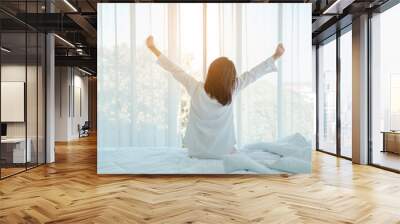 Photo of young happy woman in pajama stretching her arms and smiling while sitting on bed after sleep Wall mural