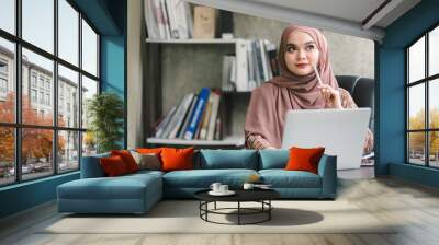 A beautiful Muslim millennial Asian businesswoman wearing a brown hijab working remotely from her home office. Wall mural