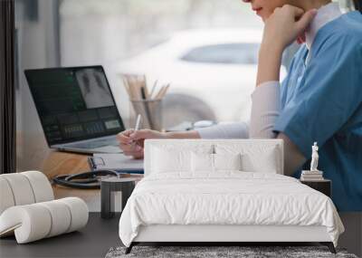 A professional female doctor is taking notes in a medical journal and filling out documents, including the patient's illness history, looking at a laptop screen dashboard. Wall mural