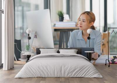 A young, happy Asian professional businesswoman is sitting at her desk, working on a tablet and desktop computer in a corporate office. Wall mural