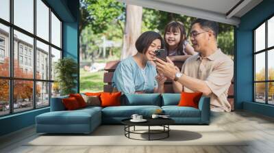 Cheerful Asian family of three having fun watching a phone together in the summer outdoors at the park. Wall mural