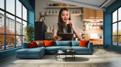 Enjoying snack while using tablet, young woman sits at wooden table in cozy kitchen. warm lighting and colorful mugs in background create cheerful atmosphere Wall mural