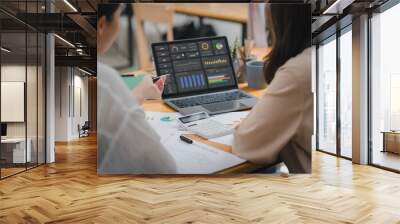 Two businesspeople or an accountant team are analyzing data charts, graphs, and a dashboard on a laptop screen in order to prepare a statistical report and discuss financial data in an office. Wall mural