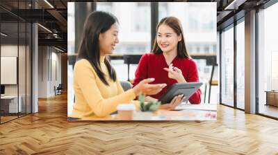 Two women engaged in friendly discussion while using tablet in modern workspace. Their expressions convey collaboration and enthusiasm, highlighting productive atmosphere. Wall mural