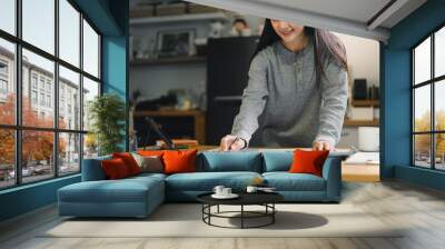 woman is working on project at wooden table, using pen to write on large sheet of paper. She appears focused and engaged in her task, surrounded by cozy home environment. Wall mural