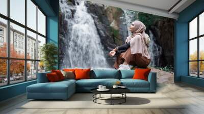 Young asian muslim tourist woman wearing brown hijab sitting on the rock in front of waterfall. Wall mural