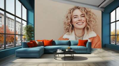 Smiling woman with curly hair in cozy autumn outfit against beige wall Wall mural