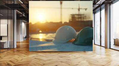 Two construction helmets rest on blueprints at building site during sunset, symbolizing teamwork and progress. cranes in background highlight ongoing development Wall mural