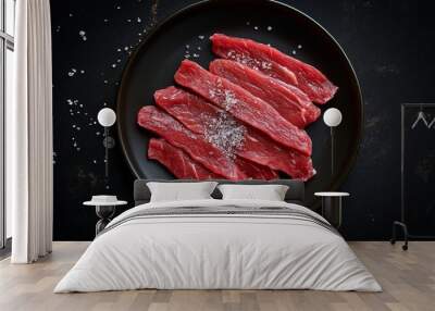 Top view of fresh raw beef slices arranged on a black plate, sprinkled with coarse sea salt, ready for cooking. Wall mural