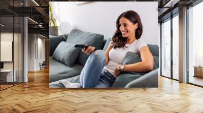 A smiling young woman sitting on the sofa and watching television. Wall mural