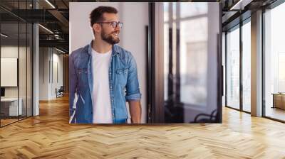 Cheerful entrepreneur in denim shirt, wearing glasses, standing in his office in the morning. Wall mural