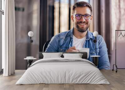 Handsome smiling young man drinking coffee sitting at office desk and looking at camera Wall mural