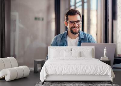 Handsome smiling young man sitting by window in office, drinking coffee while working on laptop. Wall mural