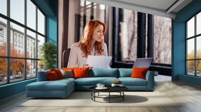 Happy smiling businesswoman in formal wear sitting at desk in modern office in front of laptop and doing paperwork Wall mural