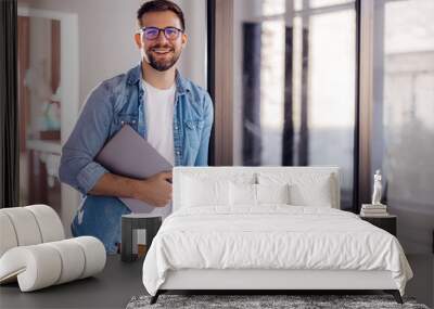 Portrait of a young modern businessman standing holding a laptop and looking at the camera with a happy smile Wall mural