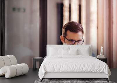 Satisfied young man with glasses sitting at a desk and doing paperwork at his workplace. Wall mural