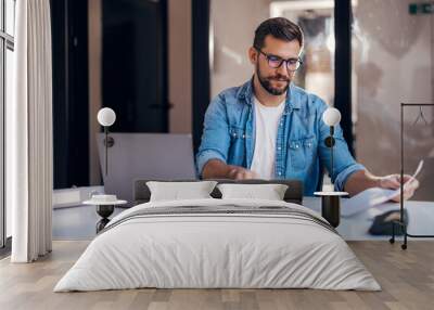 Satisfied young man with glasses sitting at a desk and doing paperwork at his workplace. Wall mural