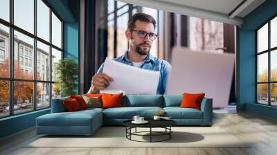 Satisfied young man with glasses sitting at a desk and doing paperwork at his workplace. Wall mural