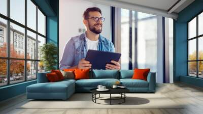 Smiling young businessman working online with digital tablet while standing by window. Wall mural