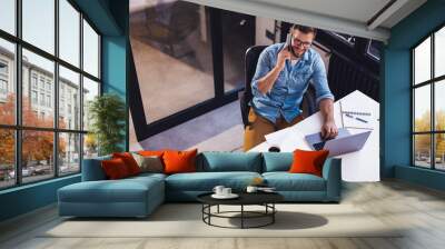 Smiling young man sitting in the office by the window and working on a laptop while talking on the phone. Wall mural