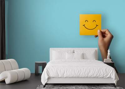 A hand holding a yellow happy smile face on blue background. Positive customer rating, good feedback and review. Happiness and world mental health day concept Wall mural