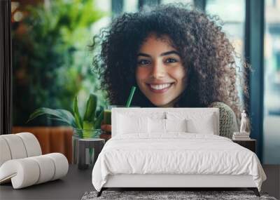 A cheerful young woman with curly hair sits at a table, holding a glass of green smoothie. Wall mural