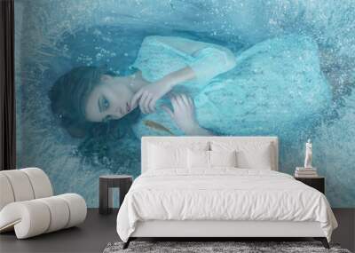 A mermaid girl in a blue vintage dress lies at the bottom of the lake. It is covered with ice edge, fish swim around it. She mysteriously looks through the ice with her finger against her lips Wall mural