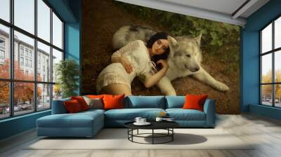 A petite girl with dark hair and soft cute face features is lying on gray-white forest wolf, doll in short white light dress resting on a big dog, a cute and attractive photo of fairy tale dream Wall mural