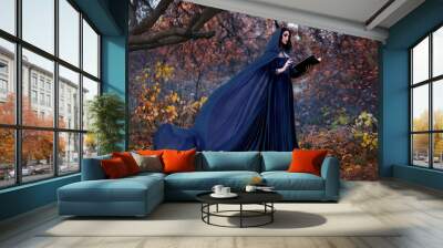 Art photography. Woman witch holds book in hands reads spell. Black clothes, cloak dress flies in wind. Fantasy girl elf in hood. Nature autumn forest, trees orange leaves. Holiday concept halloween. Wall mural