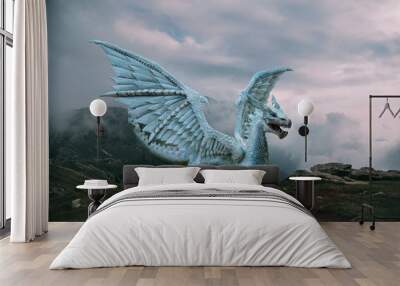 Fantasy dragon stands on the top of the mountain. A huge dangerous animal with large, sharp wings. Beautiful alpine nature background, dramatic gray-blue sky with white clouds. Wall mural