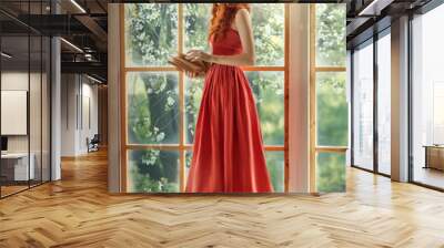 Medieval woman princess holds in hands reads book. Red-haired dreamlike girl stands on window barefoot, view summer green garden trees. Red vintage dress, ball gown. Long red curly hair, pale skin Wall mural