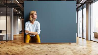 A woman in casual attire sits pensively against a gray wall, with a small plant nearby, conveying solitude and introspection. Wall mural