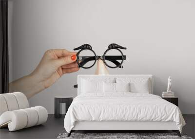 Closeup of woman hand showing fake nose and glasses, with mustache and furry eyebrows. Indoor studio shot isolated on gray background. Wall mural