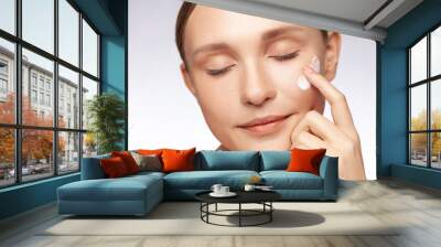 Closeup portrait of winsome beautiful woman touching her cheek applying cream grooming her facial skin, closed eyes, cosmetology concept. Indoor studio shot isolated over gray background. Wall mural