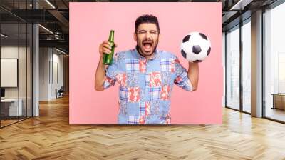 Crazy fan man wearing blue casual style shirt screaming happily, celebrating winning his favorite team, holding in hands ball and bottle with alcohol. Indoor studio shot isolated on pink background. Wall mural