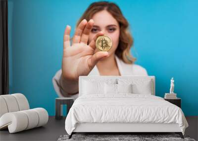 Cryptocurrency. Portrait of young woman with wavy hair in white jacket holding physical golden bitcoin, advertising of btc crypto coin, digital currency. indoor studio shot isolated on blue background Wall mural