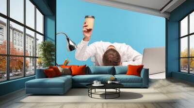 Exhausted male employee feeling fatigue lying on table and raising coffee cup, lack of energy in morning office, tired of stress problems sleeping at workplace, overwork concept. indoor studio shot Wall mural