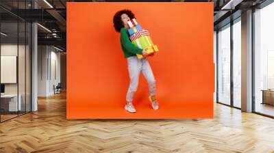 Full length portrait of woman with Afro hairstyle wearing green casual style sweater holding heavy gift boxes, carrying heavyweight presents. Indoor studio shot isolated on orange background. Wall mural