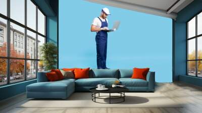 Full length professional workman in uniform and protective gloves typing on laptop keyboard, having online communication with client, reading order for repair services. indoor studio shot isolated Wall mural