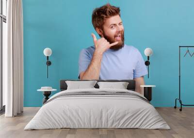 Give me a call. Portrait of positive optimistic bearded man smiling at camera and winking eye, making call me gesture with fingers, flirting. Indoor studio shot isolated on blue background. Wall mural