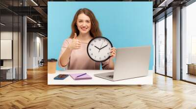 Good job, time to work! Positive happy woman, satisfied office employee sitting at workplace, holding big clock and showing thumbs up, like gesture. indoor studio shot isolated on blue background Wall mural