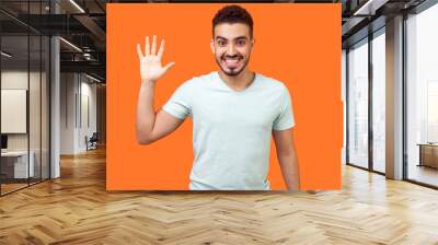 Hello! Portrait of friendly glad handsome brunette man with beard in casual white t-shirt showing hi gesture with waving hand and smiling sincerely. indoor studio shot isolated on orange background Wall mural