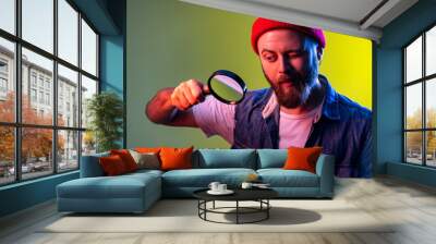 Hipster man looking through magnifying glass, spying, finding out something, exploring and inspecting, wearing beanie hat and denim vest. Indoor studio shot isolated on colorful neon light background. Wall mural